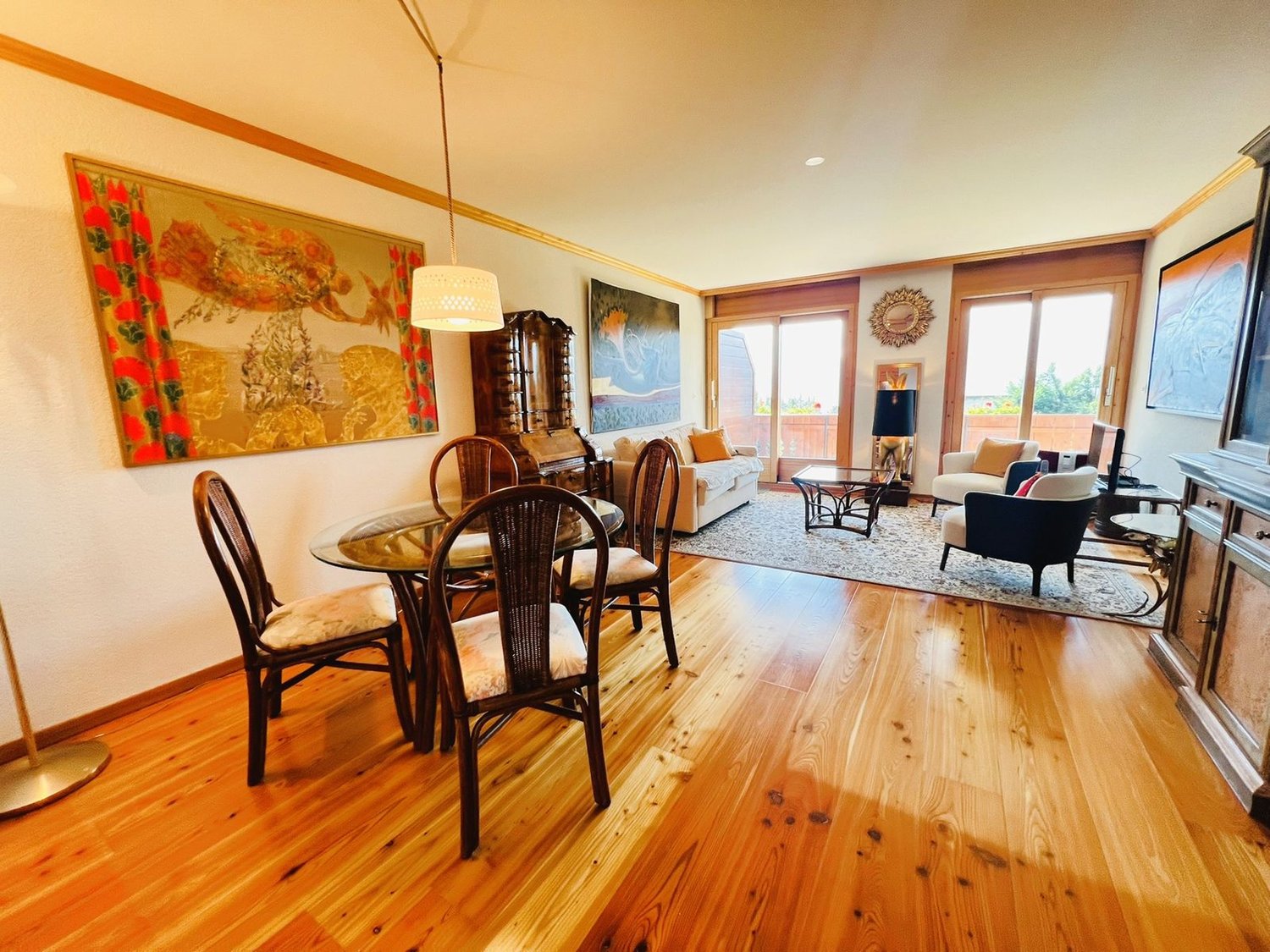 Wooden flooring, round dining table with chairs, hanging lamp, paintings on the wall, glass sliding doors, couch with pillows, side table with lamp, coffee table, rug on the floor, cabinet