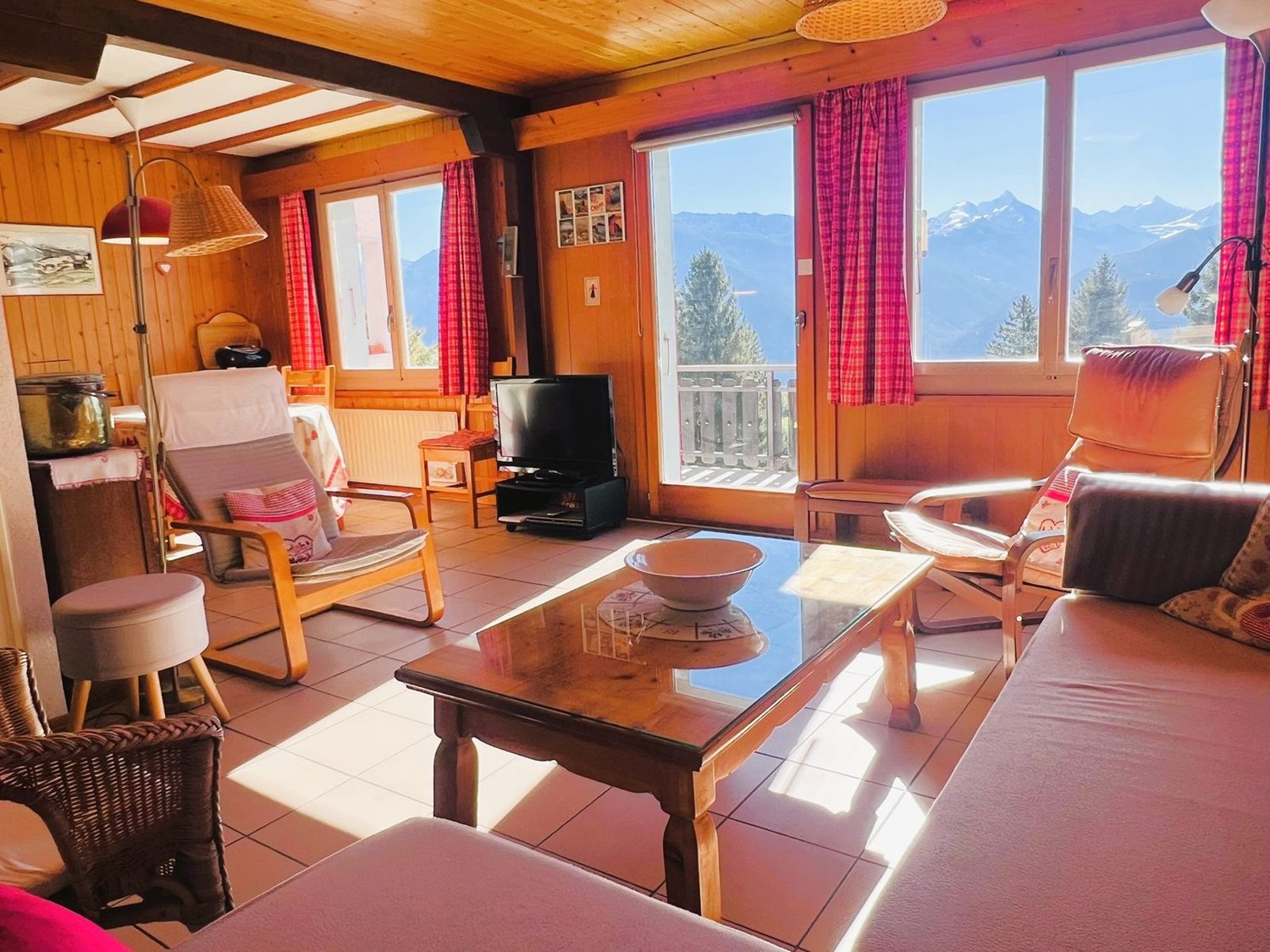 Living room with wooden walls, tiled floor, large windows, balcony with railing, couch, coffee table, armchairs, table with TV and stand, floor lamp, stool, framed picture, picture on the wall