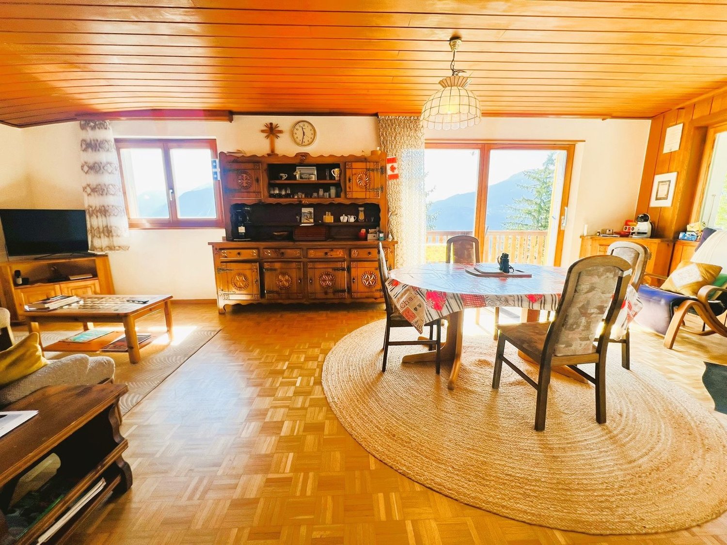 wooden floor, wooden ceiling, dining table, chairs, round rug, wooden cabinet, TV, clock, windows, mountains view