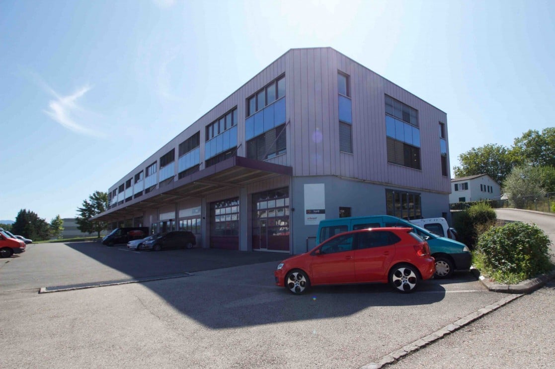 A multi-story commercial building with a modern, angular design featuring large windows. The building has a parking lot in front with several cars parked. The surroundings include some trees and greenery.