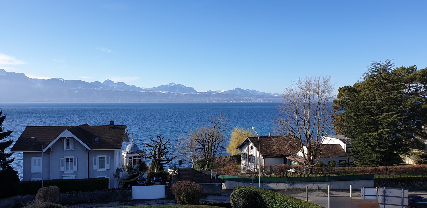 The image shows a scenic view of a lakeside town surrounded by mountains. There are several white houses with red roofs visible, set against the backdrop of the blue lake and distant mountain range. The foreground includes some trees and landscaping eleme