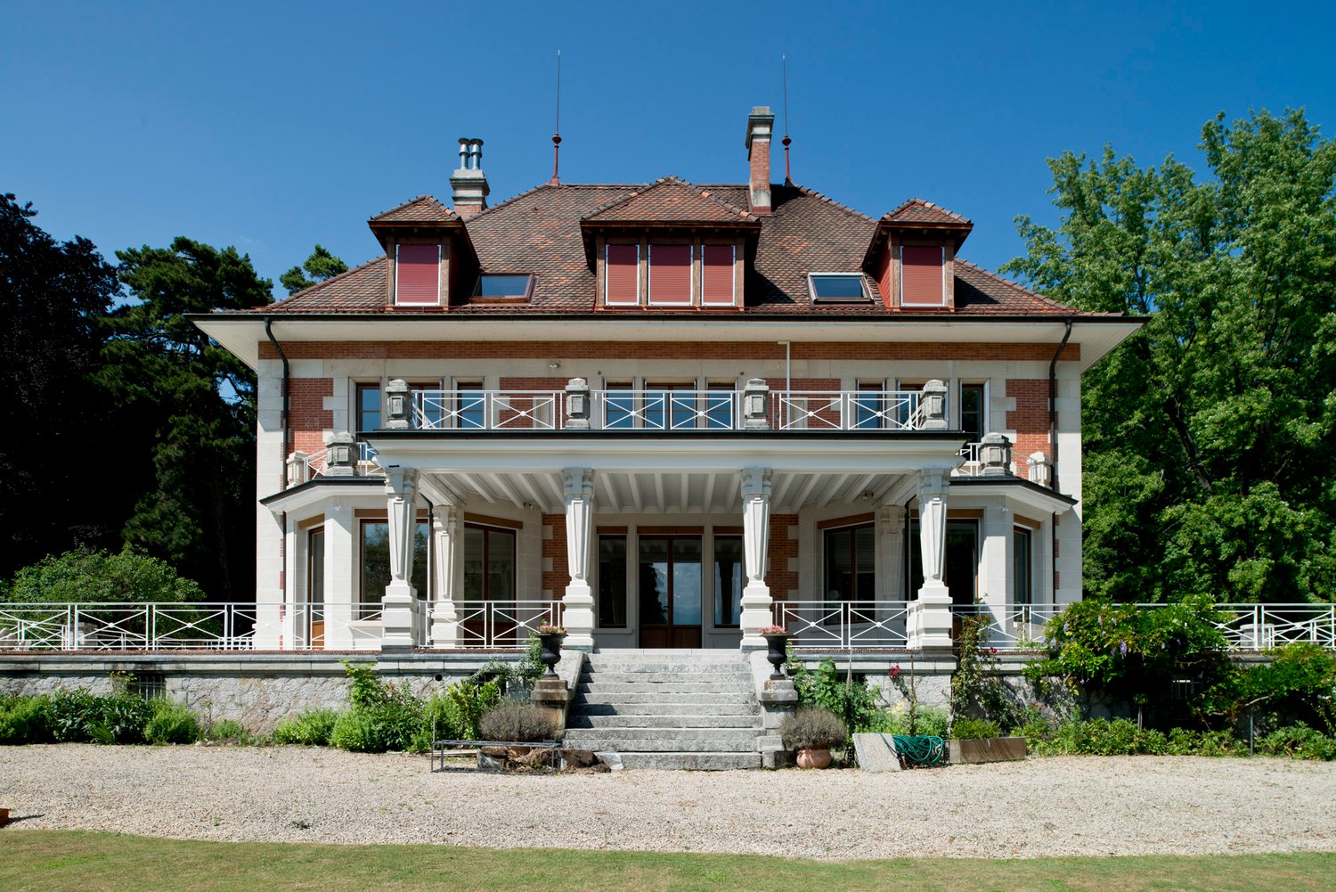 Raised ground floor, tiled roof, brown brick, white exterior, large balcony with railings, staircases, stone porch, windows, plants