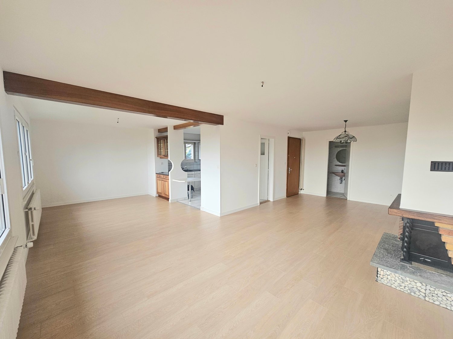 Empty room, fireplace, wooden floors, white walls, windows, wooden beam on the ceiling, wooden kitchen cabinets, radiators.