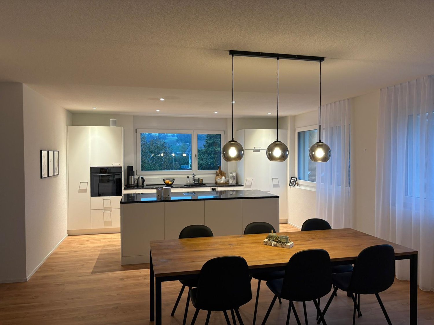 Open-plan kitchen with dining area, large window, modern lamps, wooden floor, black furniture.