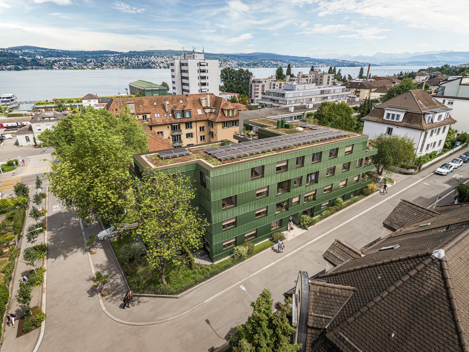 Aerial view of an apartment building with green facade, solar panels on the roof, surrounded by greenery, several houses, lake view, street with cars and pedestrians
