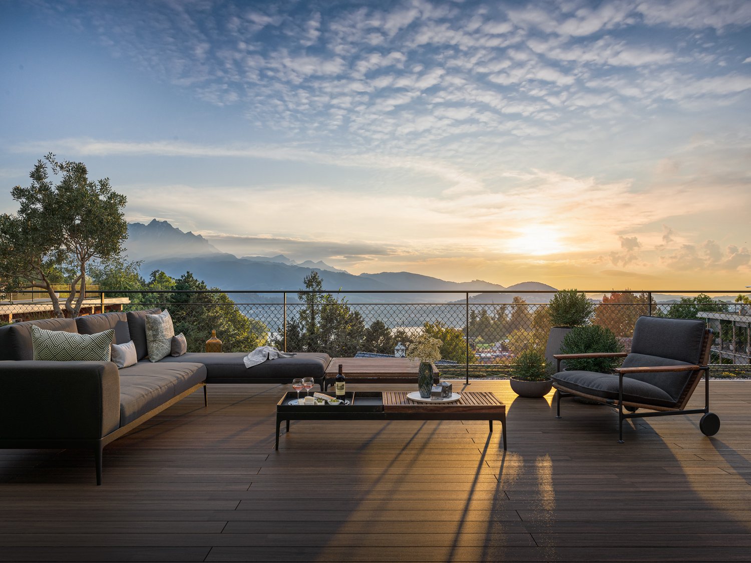 Wooden deck with railing, outdoor sofa, table with wine bottles and glasses, wooden coffee table, potted plants, sunset view