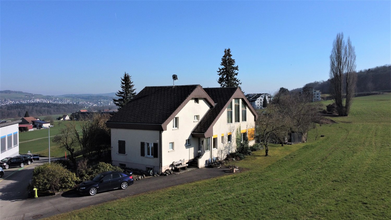 Two-story house, white exterior, black roof, surrounded by greenery, several cars parked