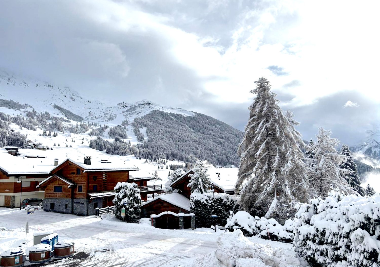 Snowy mountain landscape with several wooden chalets, trees covered in snow, and a wide snowy mountain in the background