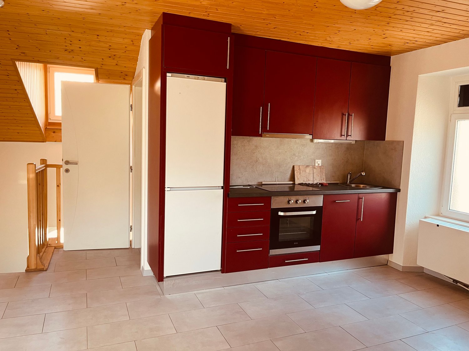 The image shows a modern kitchen with burgundy-colored cabinets, a built-in oven, a refrigerator, and a tile floor. The kitchen appears to be part of a larger living space with a wooden ceiling and a doorway leading to another room.
