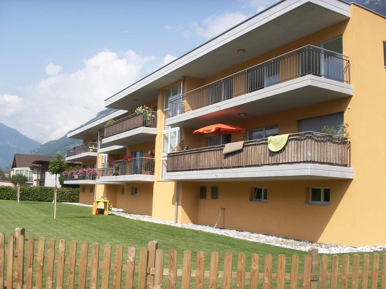 Three-story building with balconies, yellow exterior, large green lawn, wooden fence, mountains in the background