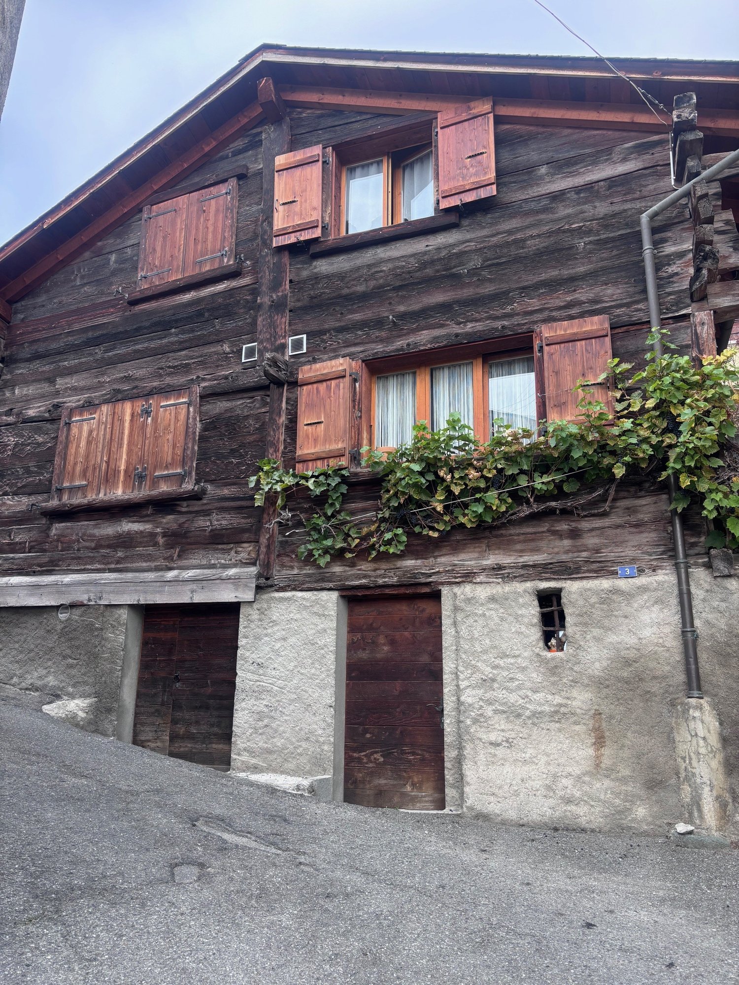 a wooden house with multiple windows and brown doors, there is a small balcony with vines at the second floor, there is a chimney attached to the wall