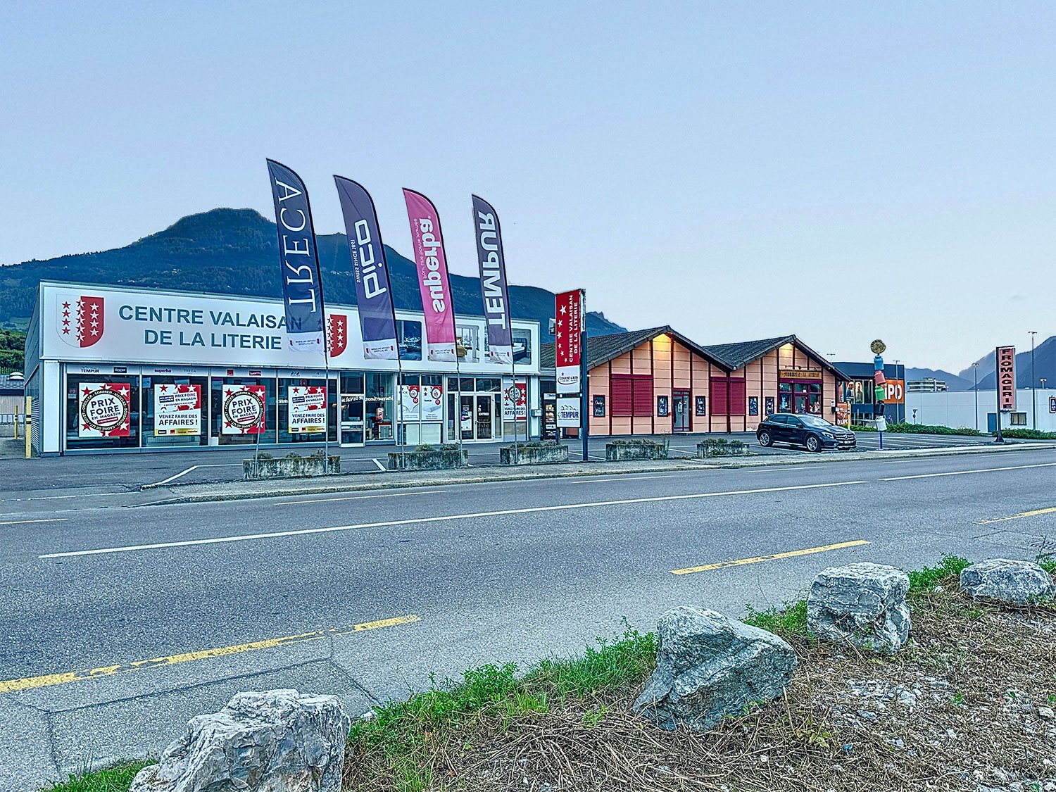 A commercial building with a car parked outside, flags on poles, large windows, red doors, mountains in the background.