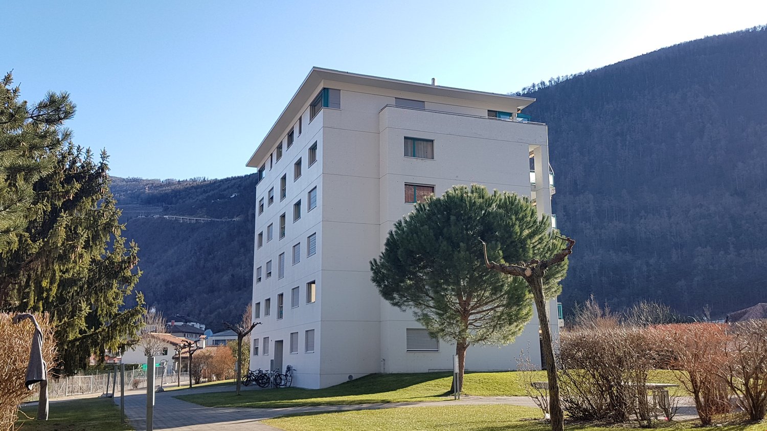 A tall apartment building, painted white, featuring multiple windows, a sloping roof, and bicycles parked on the front