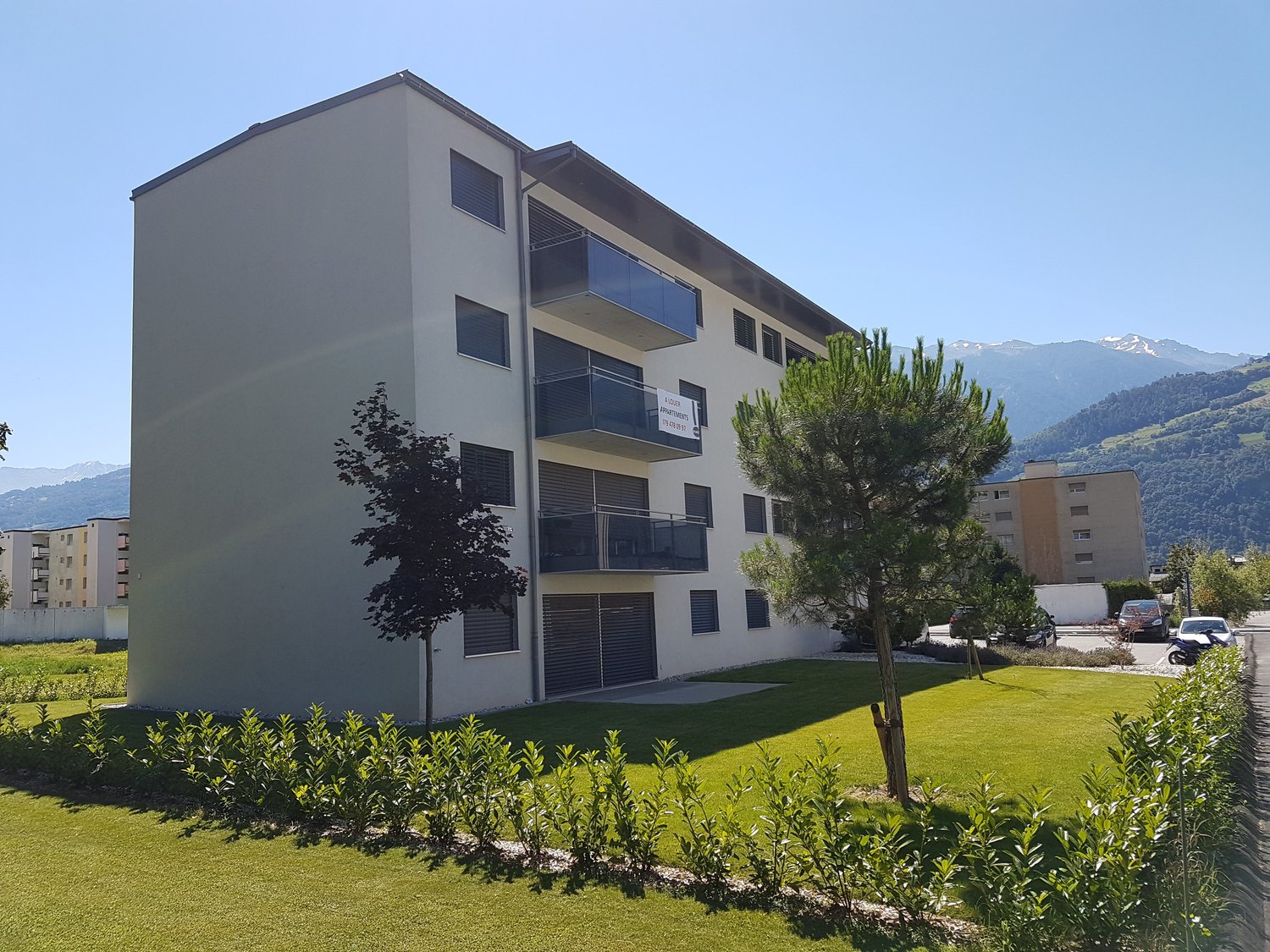 4 story building, white wall, windows with balcony, two cars parked, tree and grass, mountain view