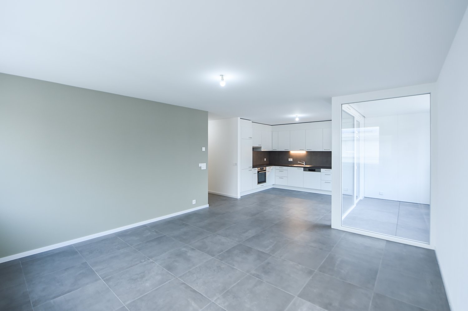 Large empty room, tiled floor, white walls, kitchen with appliances and open door