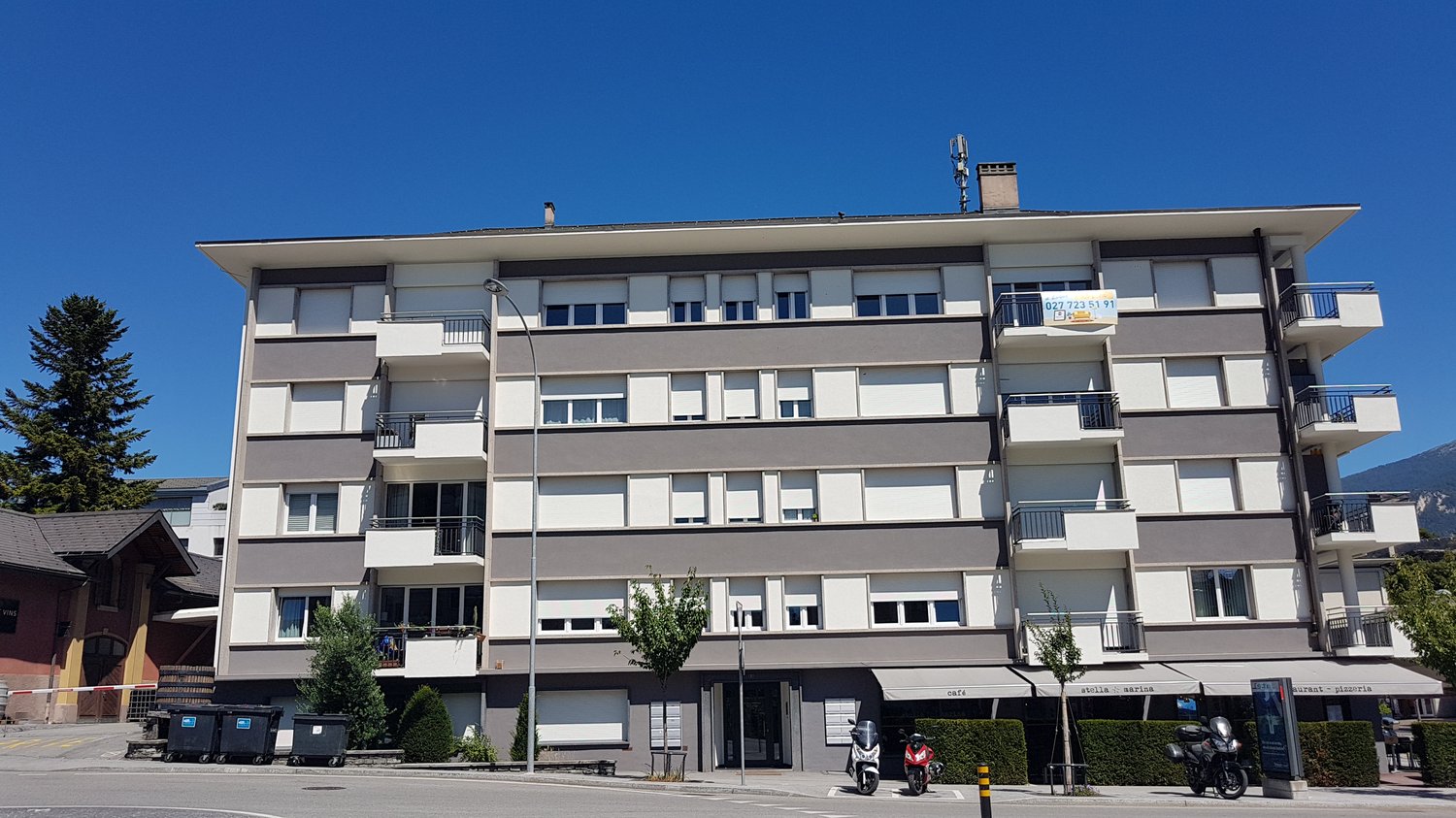 Apartment building, gray and white, multiple balconies, several floors, trees and bushes in front, two scooters parked