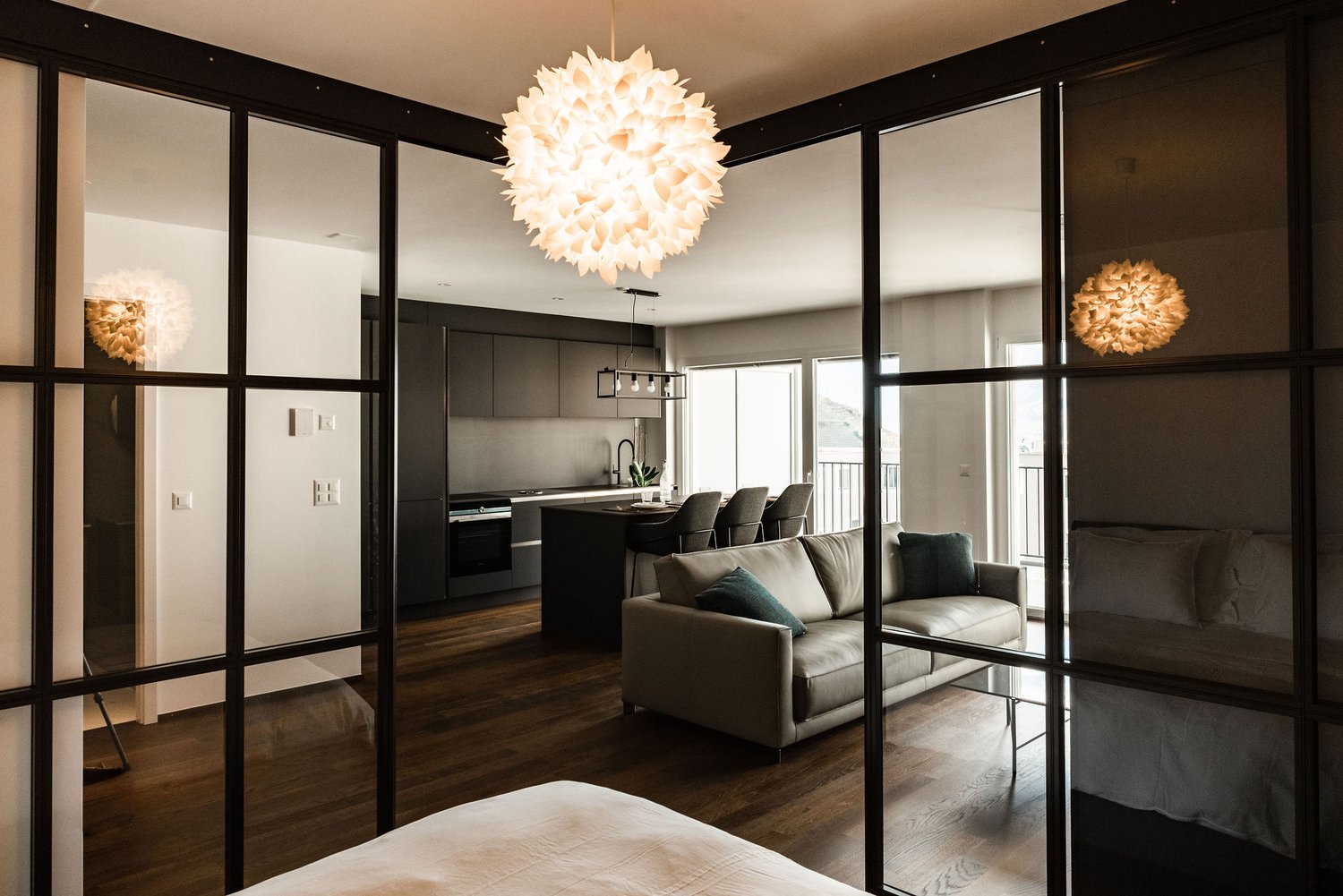 modern living room with glass walls, wooden floor, gray couch, coffee table, kitchen with island, dining table, balcony