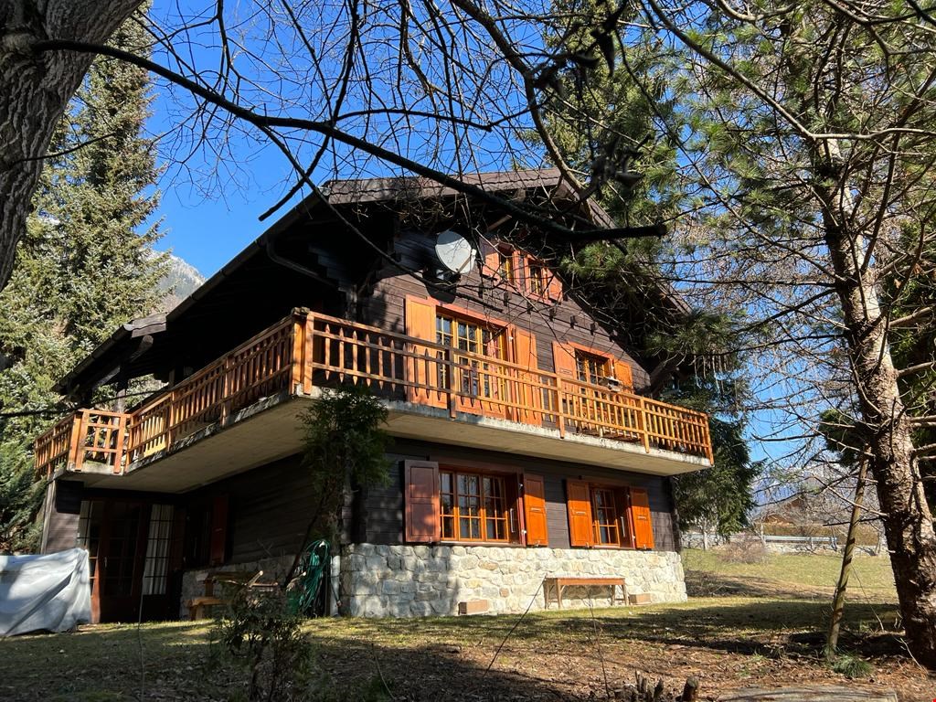Two-story wooden chalet-style house with a balcony, surrounded by trees and a grassy area