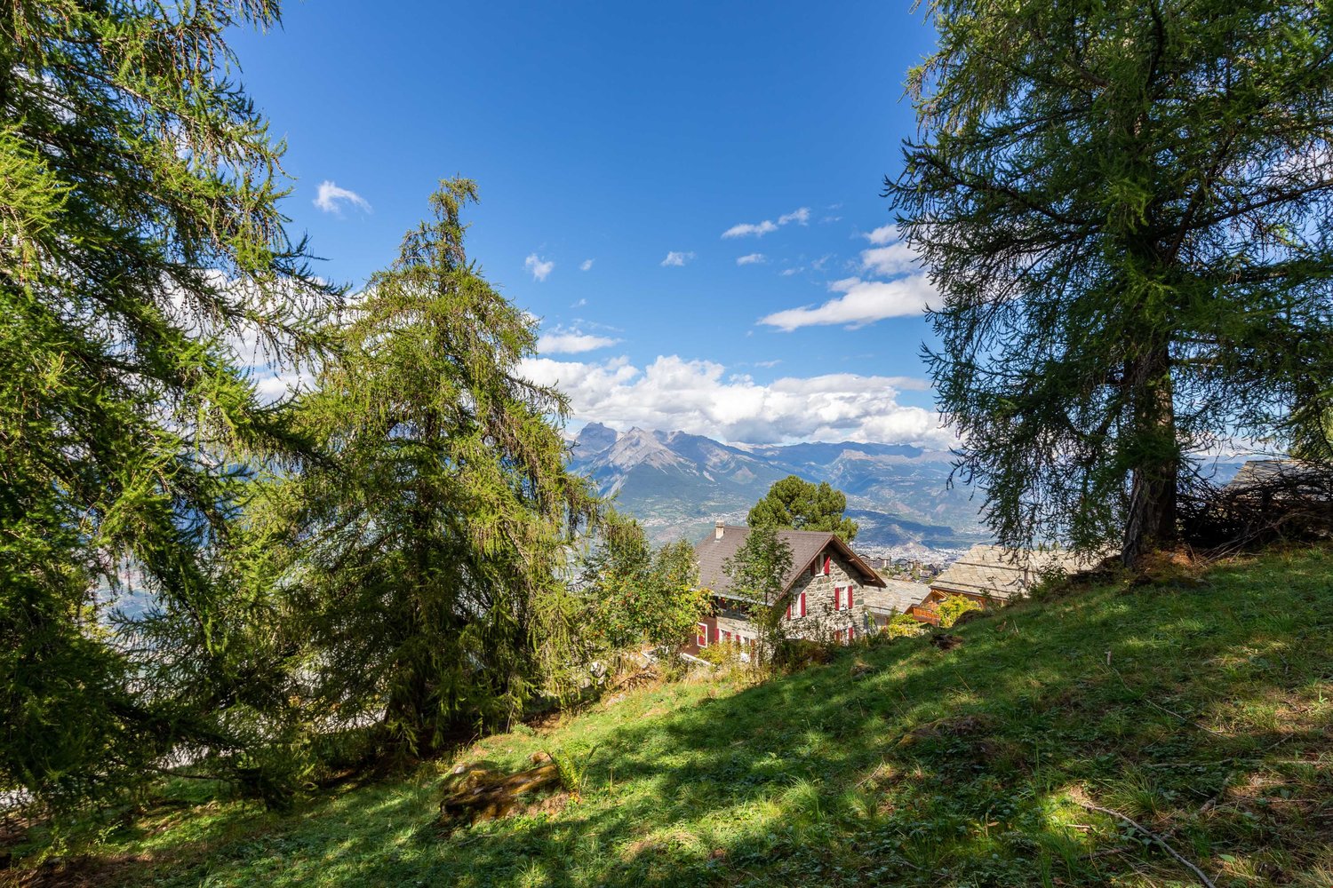Traditional stone house with red shutters, surrounded by a lush green landscape with a stunning mountain view.