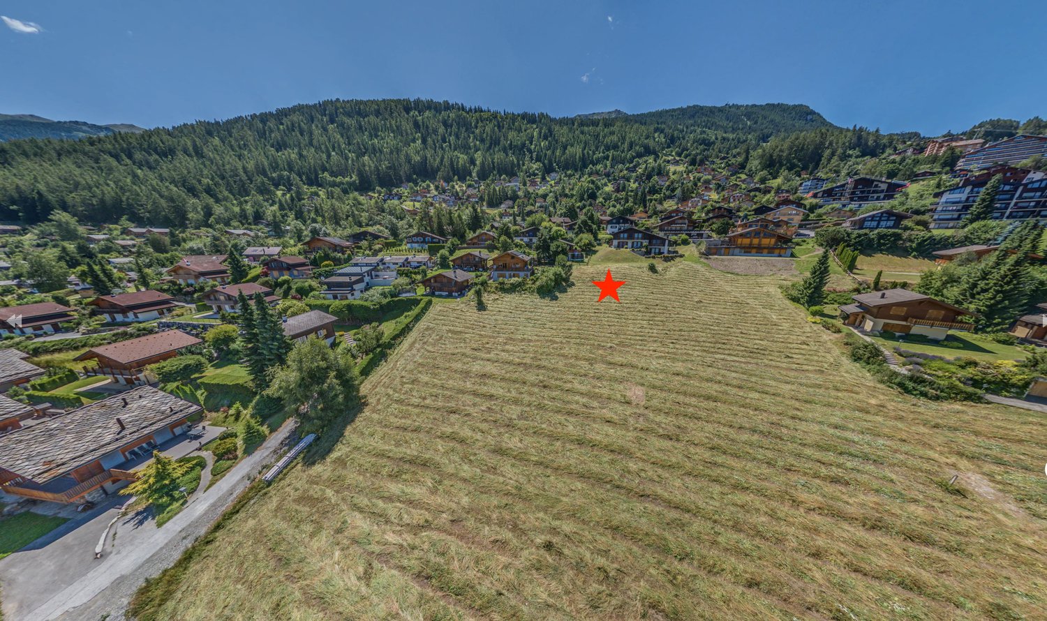 Wide field marked with a red star, surrounded by alpine houses and green trees, distant mountain range
