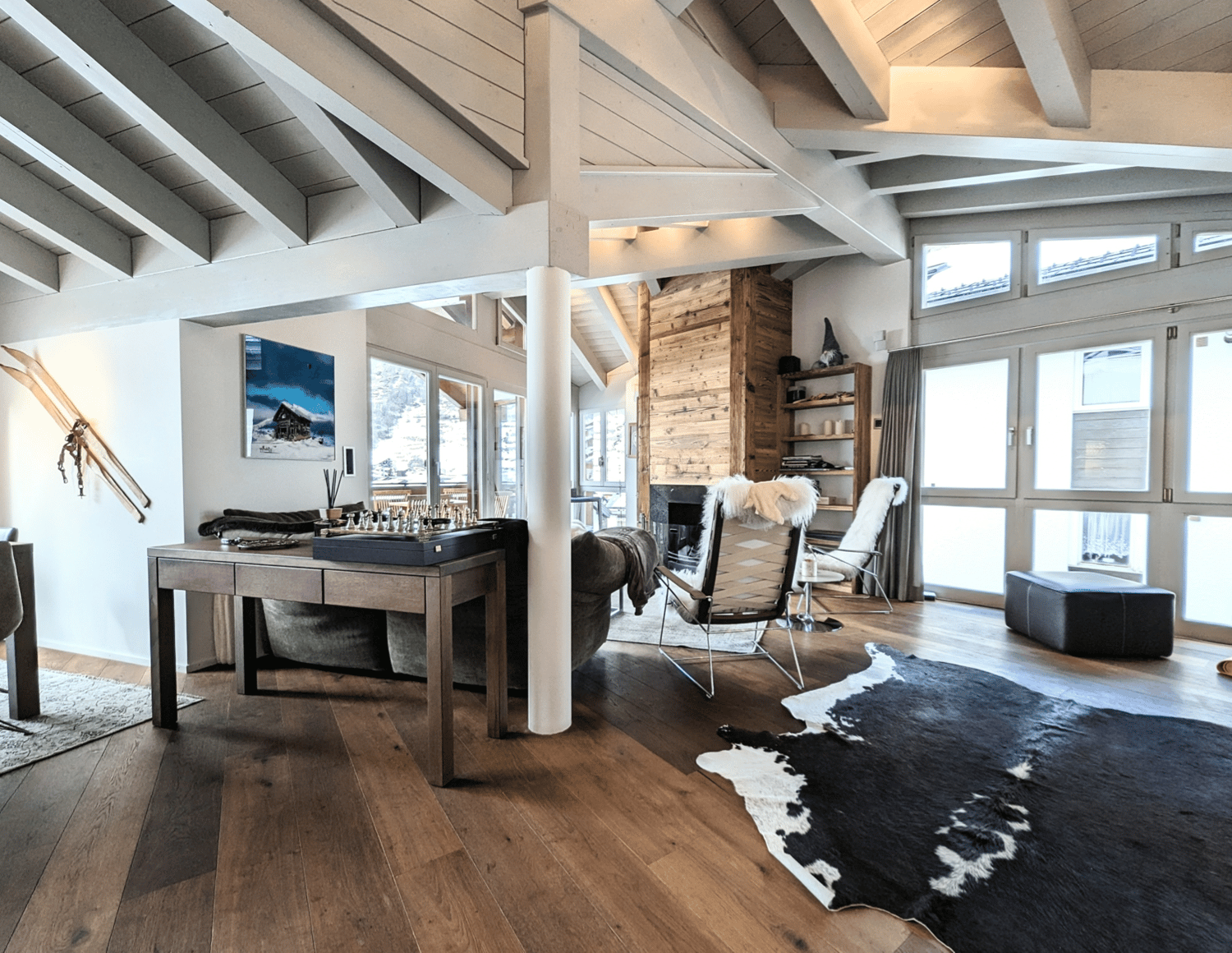 beamed ceiling, wooden floor, fireplace, sofa, coffee table, chess board, two armchairs, carpet, desk, picture on wall