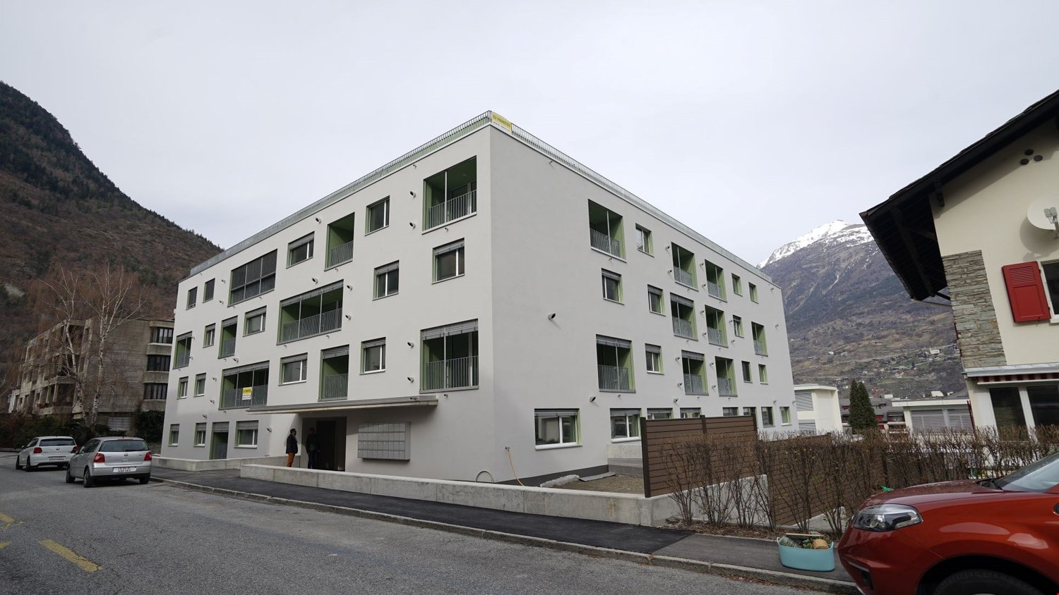 Multi-story apartment building with gray exterior, balconies, and windows. The building is situated in a mountainous area with other buildings and vehicles visible in the surroundings.