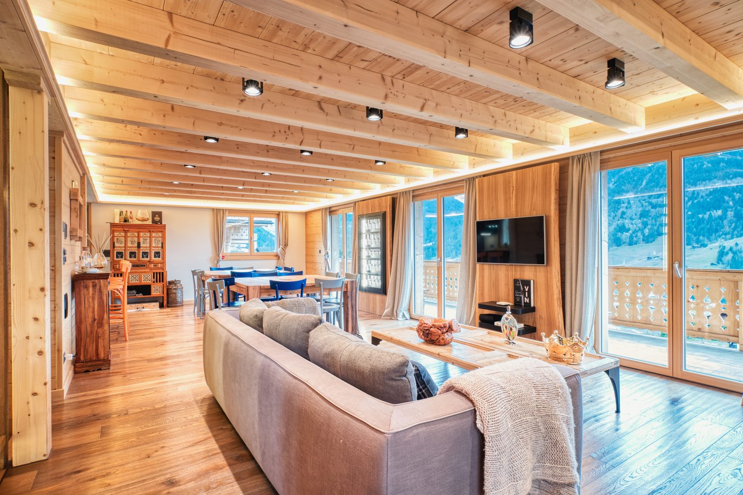 Open-plan living area with wooden beams, large sofa, center table, wooden floors, balcony, large windows