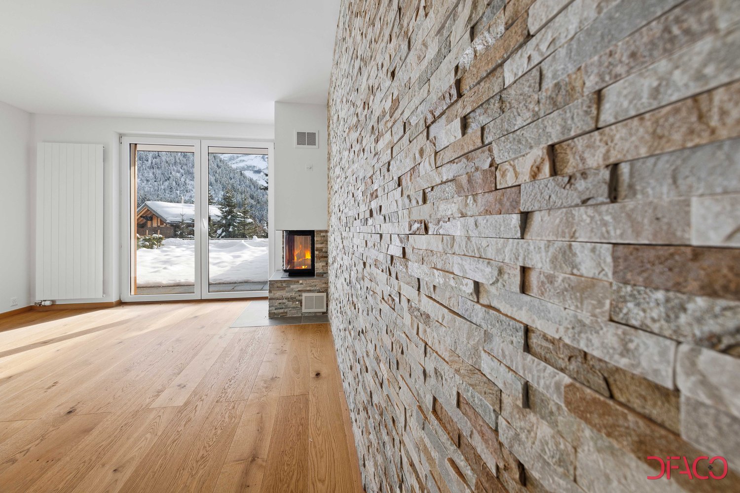Spacious living room with a stone fireplace, large windows overlooking a snowy landscape, and hardwood flooring.