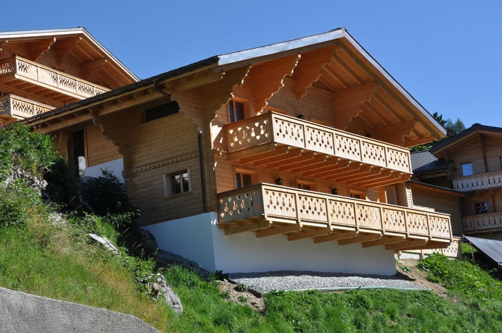 2-story wooden chalet-style house with decorative balconies, located on a hillside with a grassy slope and surrounded by trees
