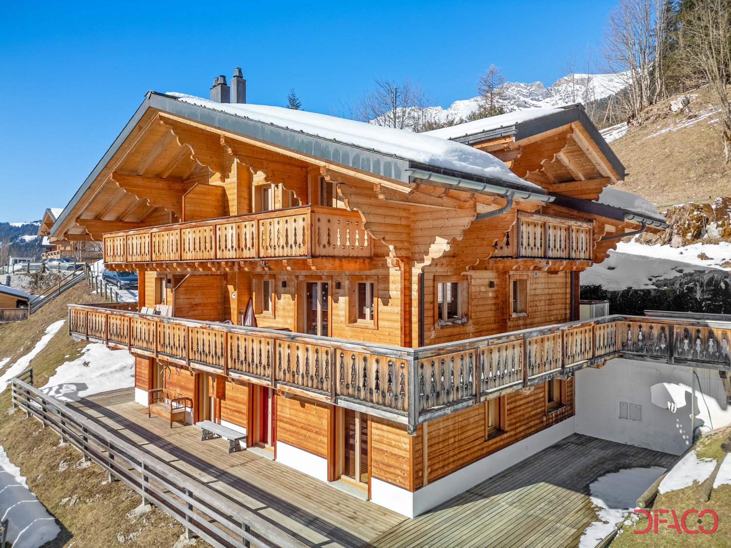 Two-story wooden chalet with a sloped roof, covered balconies, and a snowy mountain backdrop
