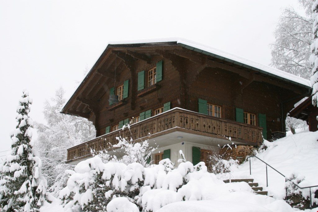 2-story wooden chalet-style house with a balcony, covered in snow in a winter landscape