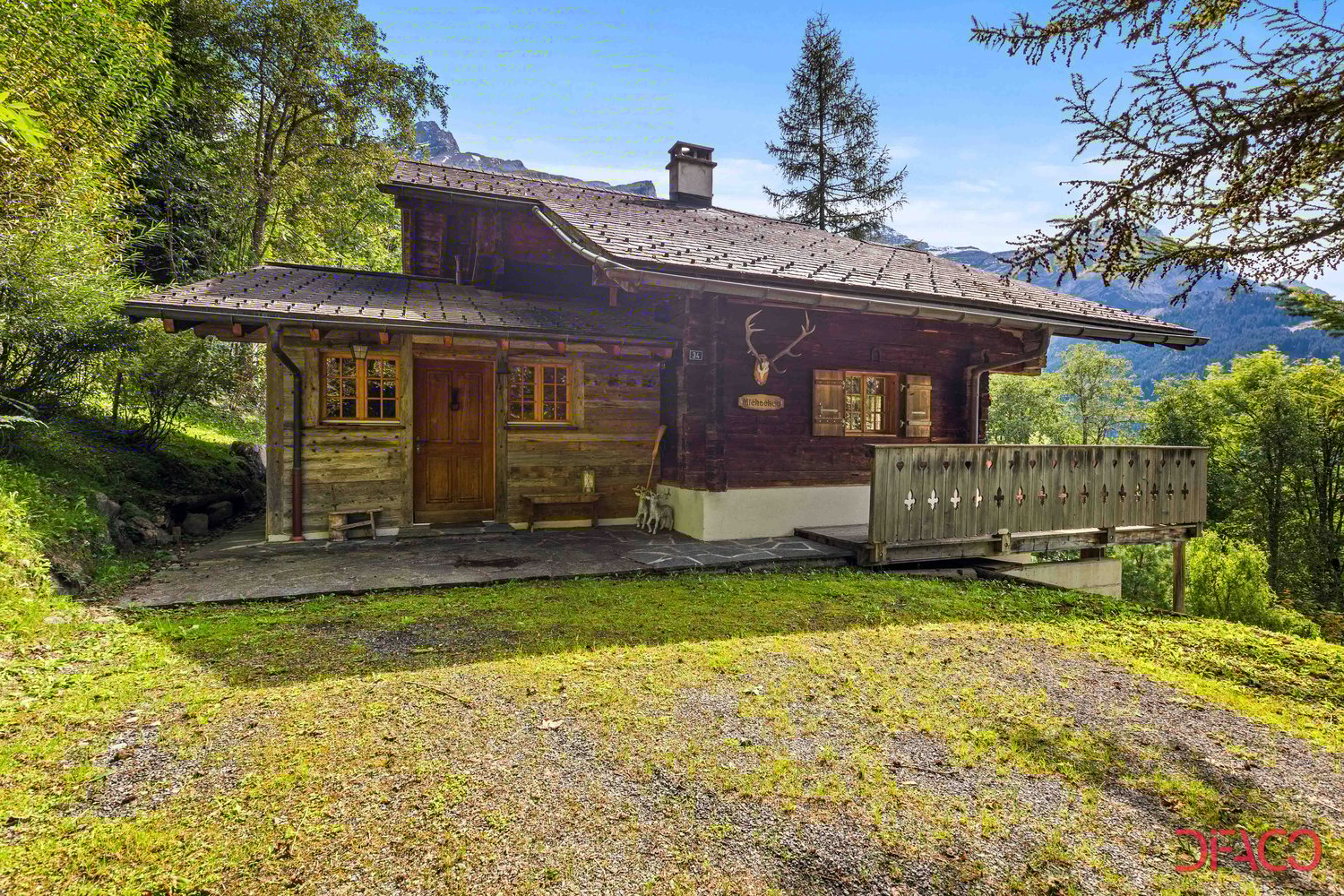 Traditional alpine-style wooden cabin with a sloped roof, surrounded by lush greenery and mountains in the background. The cabin has a wooden balcony or terrace, and a chimney on the roof.