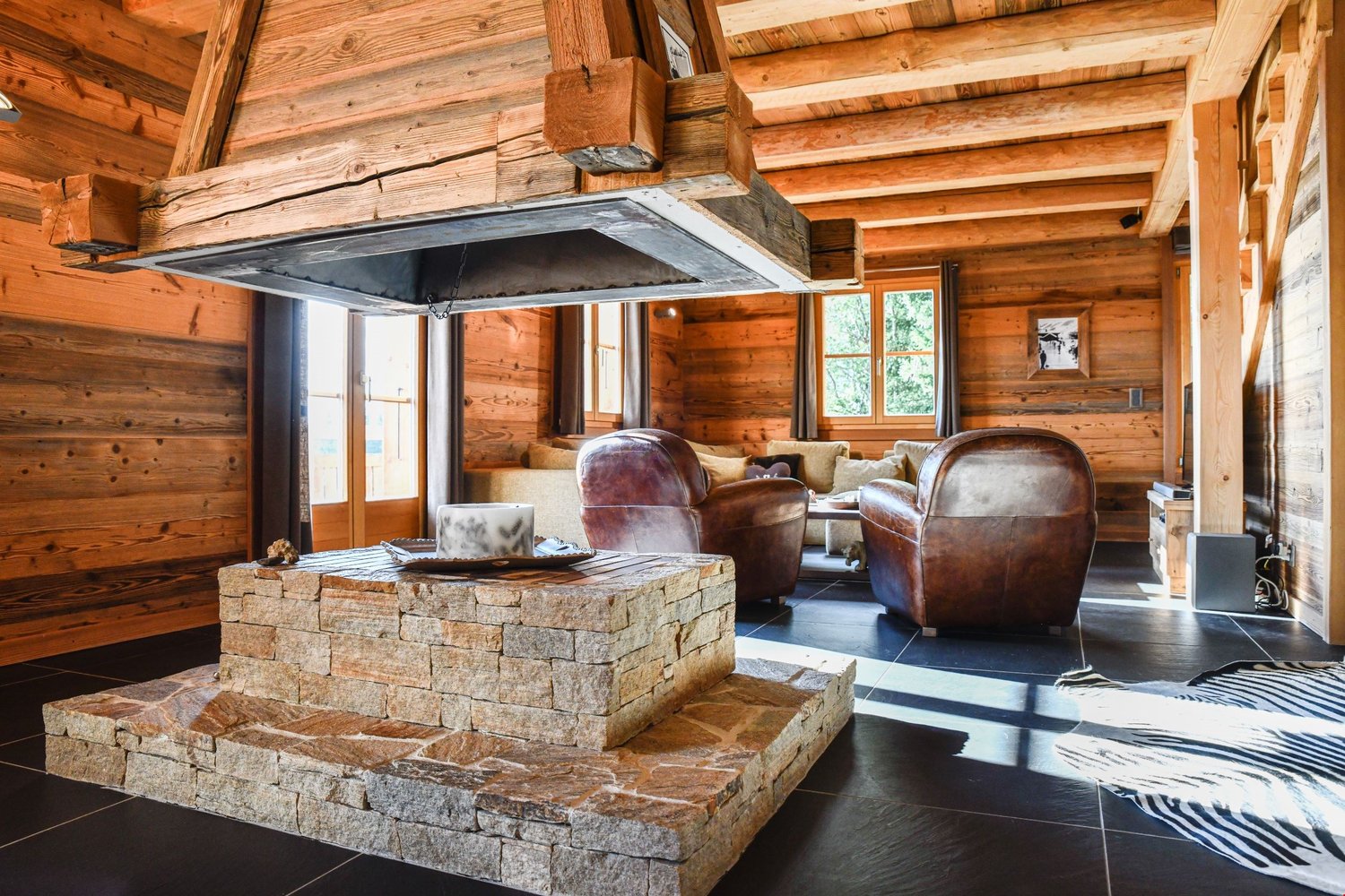 Rustic log cabin interior with wooden walls and ceiling, stone fireplace, leather armchairs, and a coffee table on a tiled floor