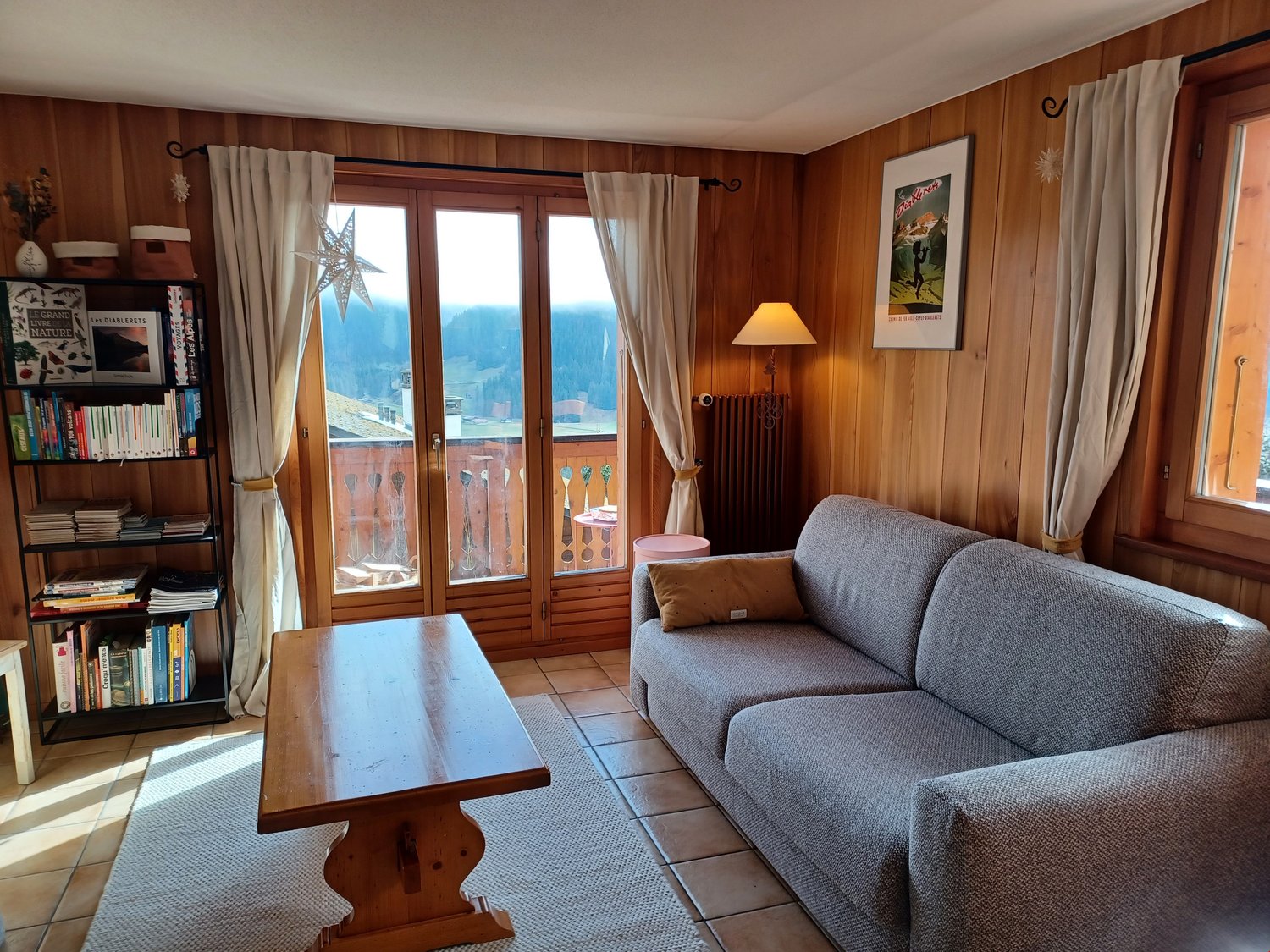 Cozy living room with wooden paneled walls, large windows with a balcony/terrace view, a gray sofa, a wooden coffee table, and a bookshelf with various books and decor items.