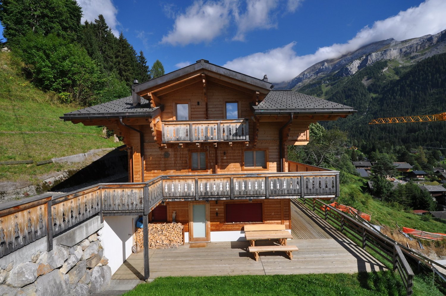 2-story wooden chalet-style house with a balcony, wooden deck, and parking space. The house is surrounded by lush green trees and mountains in the background.