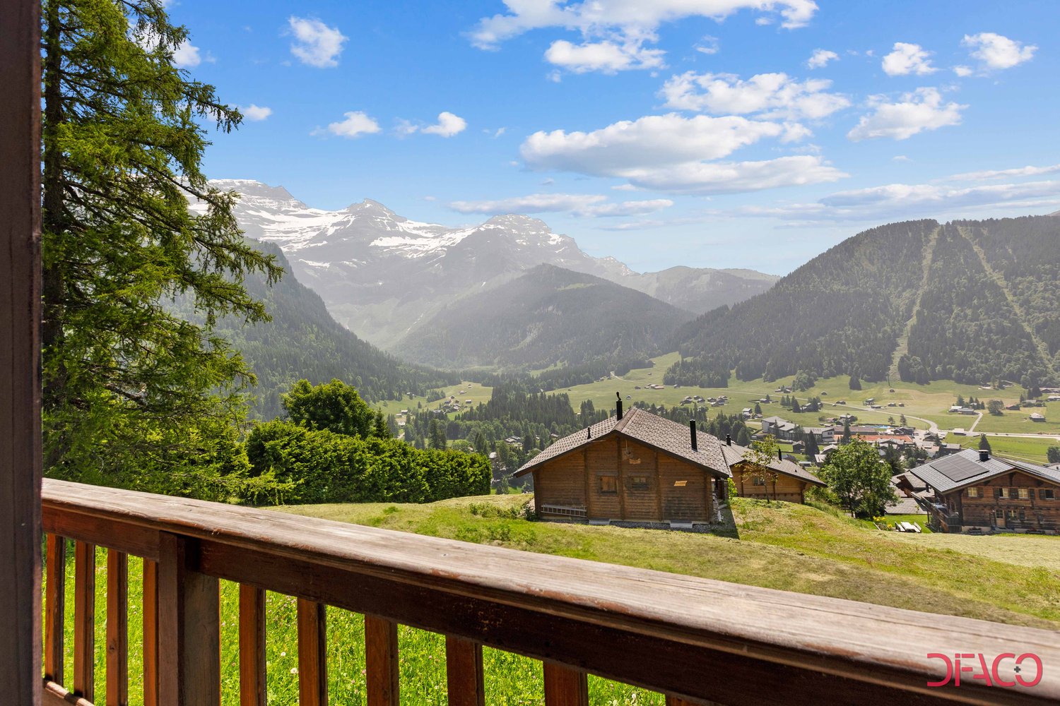 The image shows a scenic mountain landscape with snow-capped peaks in the distance, surrounded by lush green forests and meadows. In the foreground, there are several wooden cabins or chalets nestled in the valley, with a wooden deck or balcony offering a