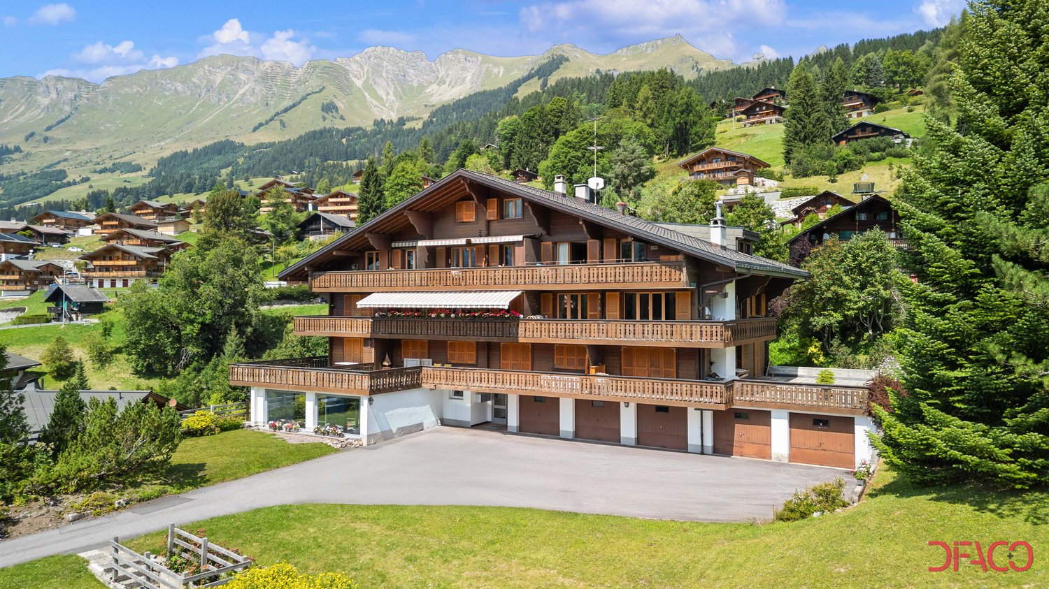 Large wooden chalet, multiple balconies, garage, set in a mountainous area with green slopes and pine trees.