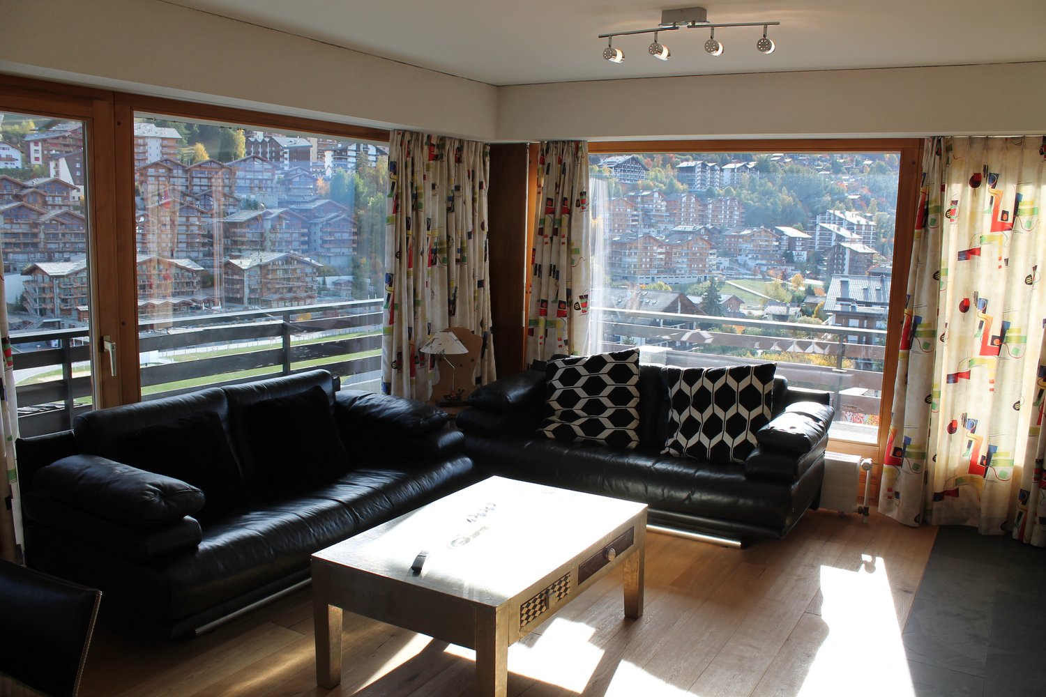 Spacious living room with large windows offering a scenic view of the surrounding buildings and landscape. The room features a black leather sofa, a wooden coffee table, and colorful patterned curtains. The lighting is provided by a track lighting fixture