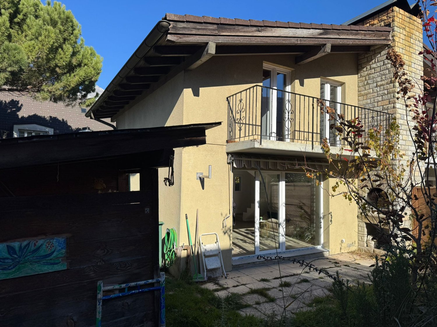 Two-story house, beige exterior, brick wall on the right side, glass door and windows, balcony with railings, a garden hose and ladder are placed outside