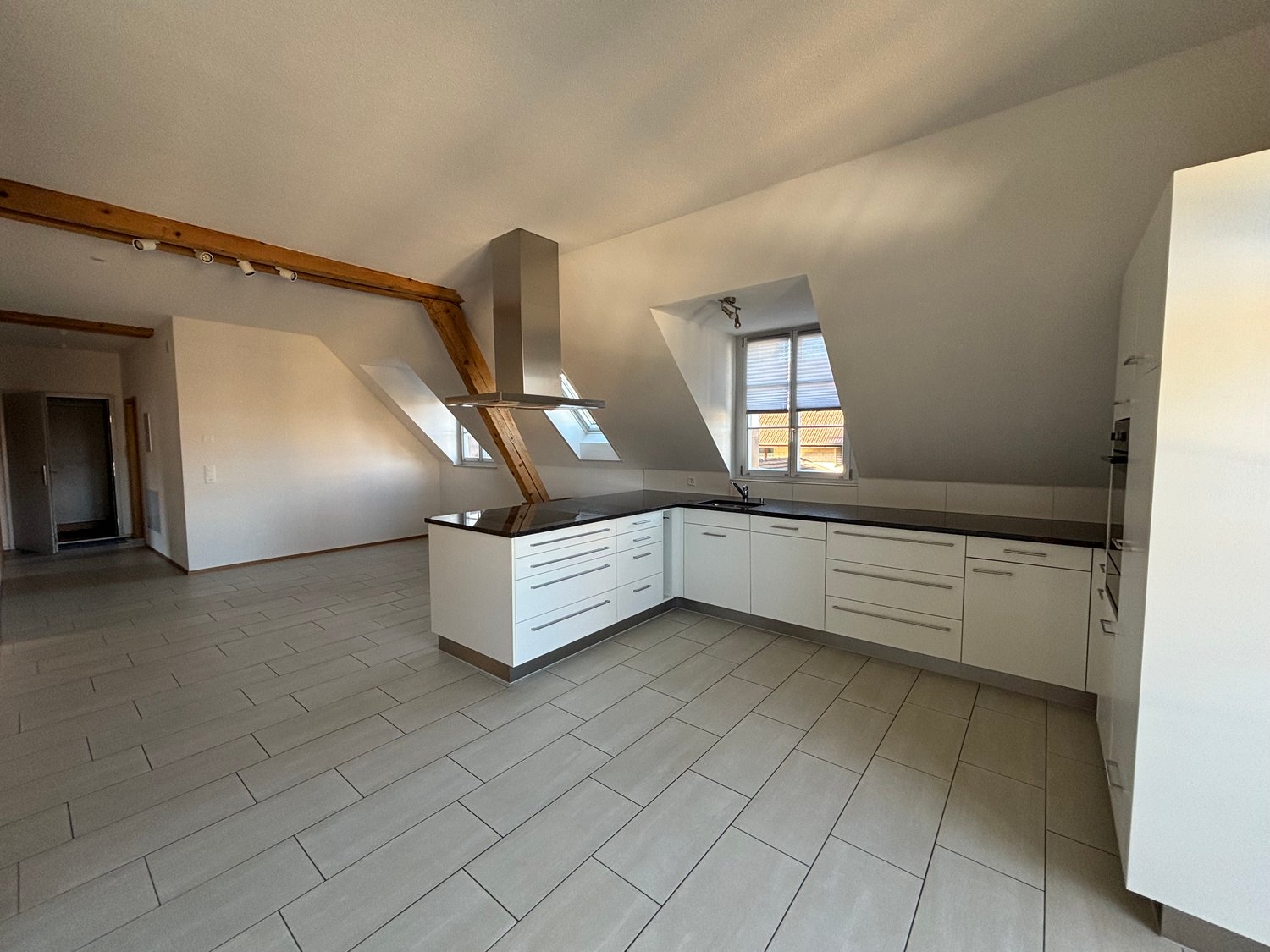 Open plan kitchen, modern cabinets, stainless steel range hood, two dormer windows, tiled flooring, ceiling beams, white walls