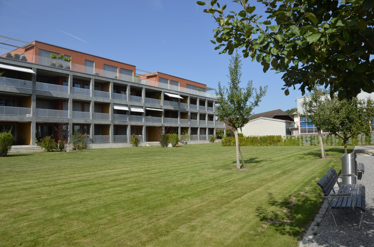 3 story apartment building, balconies, paved area with bench, green lawns, and shrubs