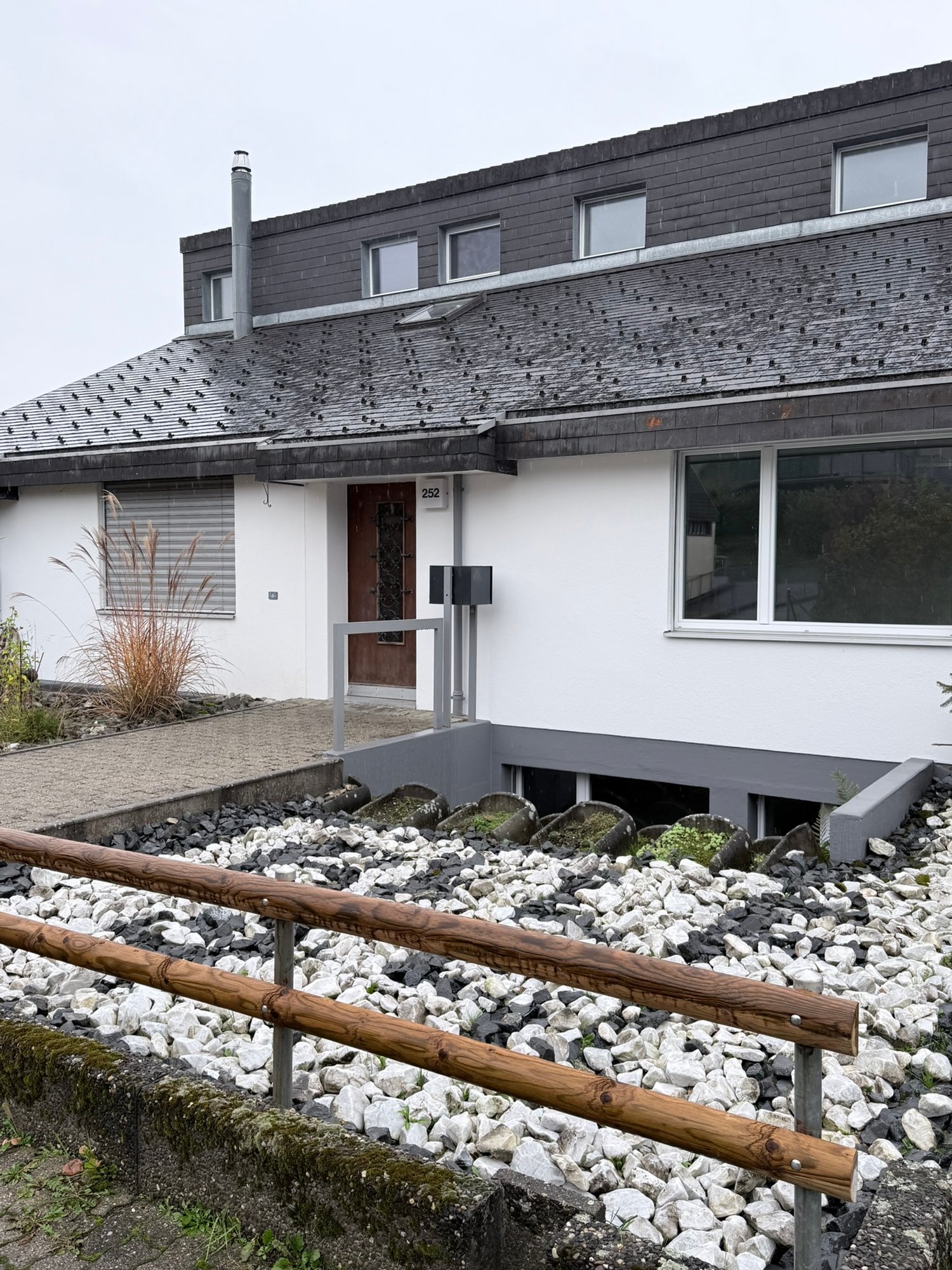 Raised ground floor, brick roof, white walls, wooden door, multiple windows, gravels, plants, number 252