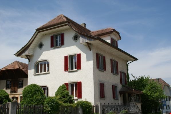 Two-story house, white exterior, red shutters, brown roof, chimney, surrounded by greenery