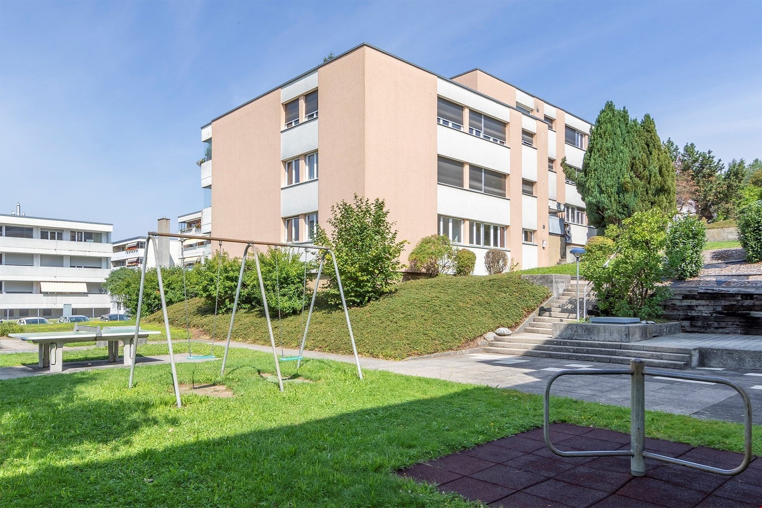 A multistory apartment building with glass windows, exterior staircase, green bushes and grass.