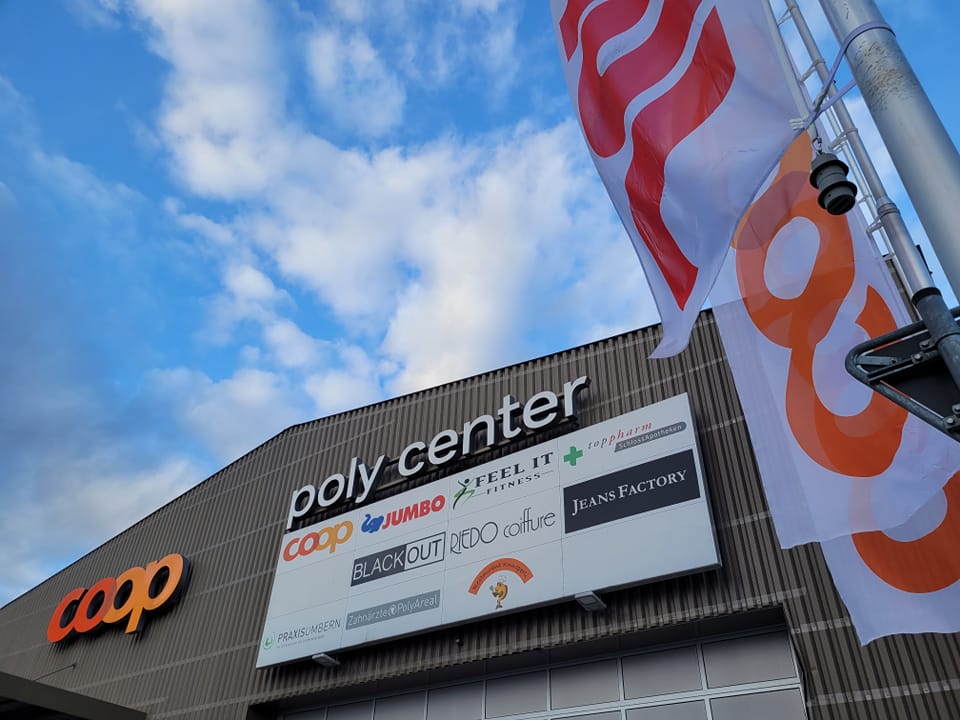 Commercial building with a large sign reading 'poly center', multiple logos on the sign, and flags on poles