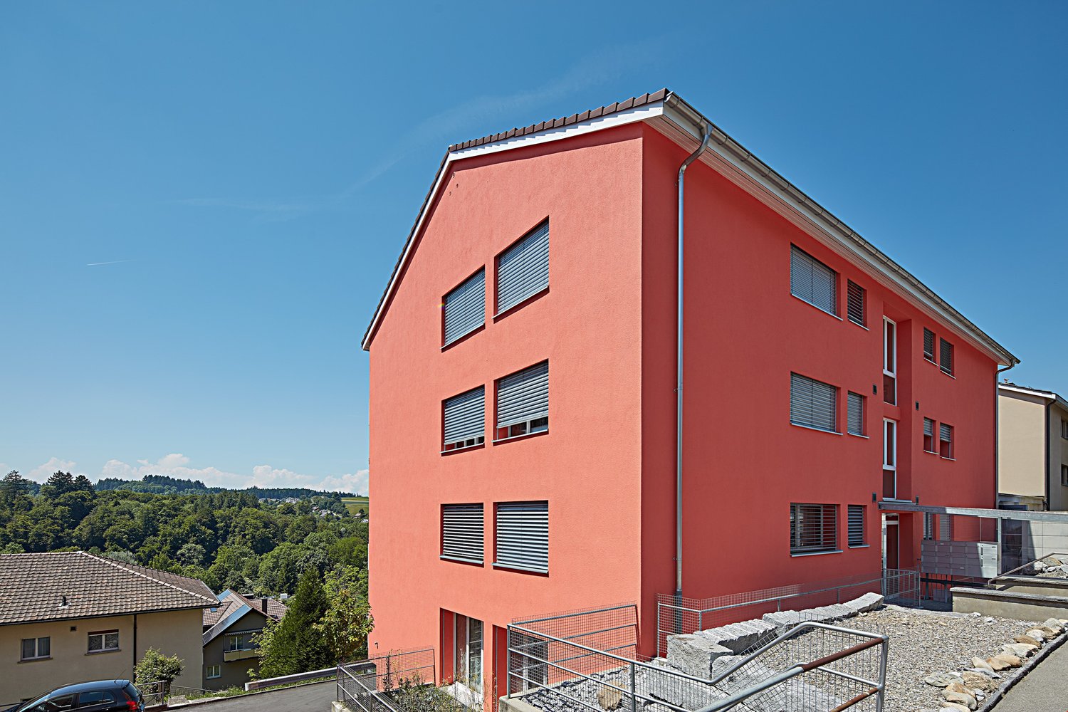 Modern apartment building, red exterior, multiple windows, metal railings, gravel pathway, nearby car