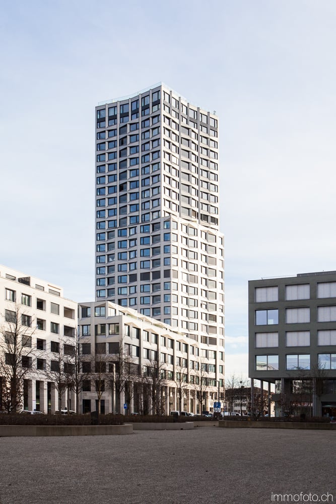High-rise building with modern design, multiple floors, many glass windows, white exterior, located in a city with other buildings and trees