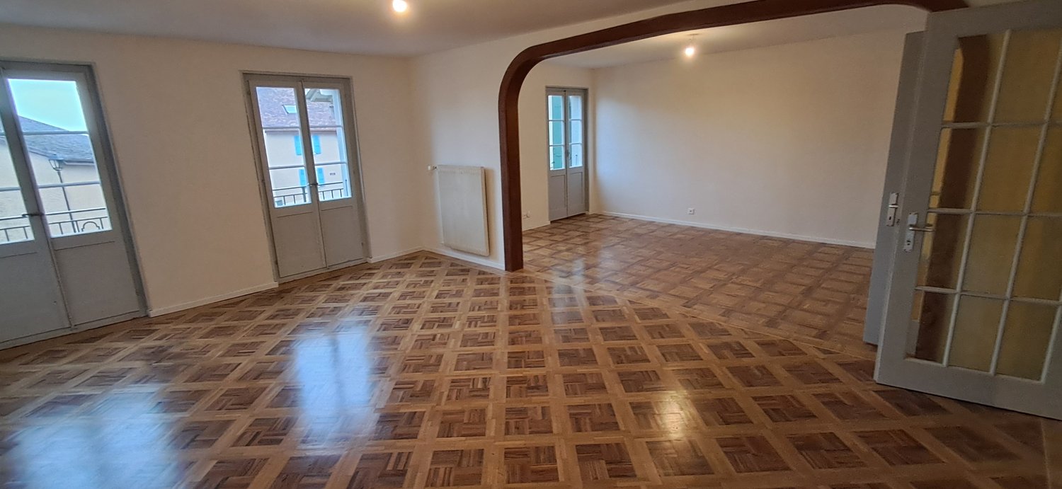 Spacious living room with hardwood parquet flooring, white walls, large windows, and a wooden beam structure overhead.
