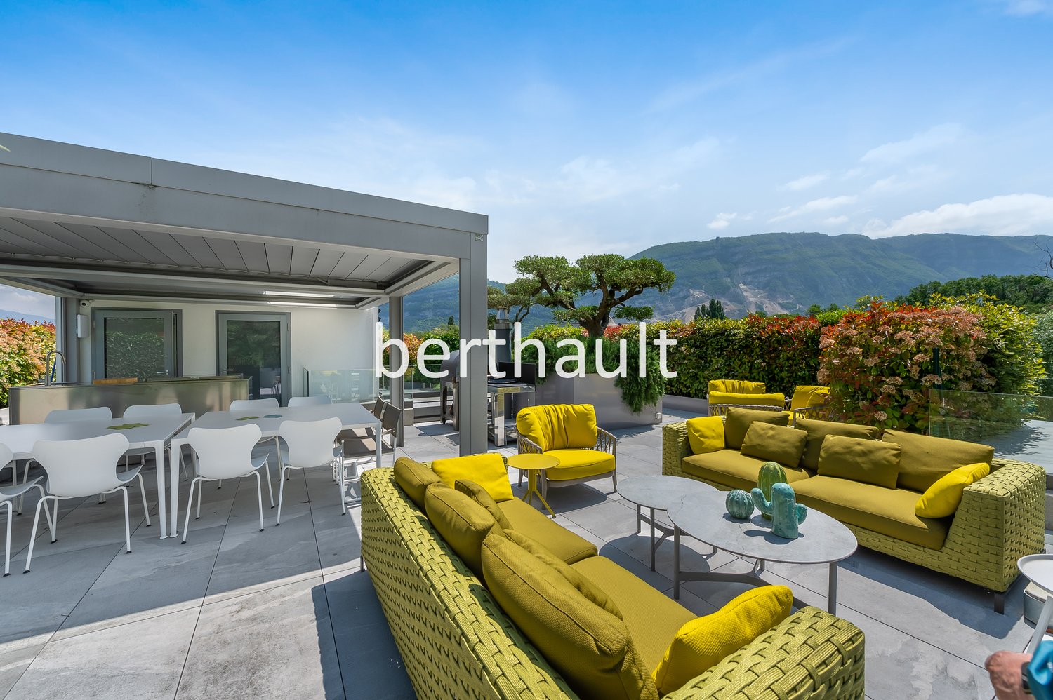 Spacious outdoor patio with modern furniture, including yellow wicker sofas and chairs. The patio overlooks a scenic mountain landscape with lush greenery. There is a covered dining area with a table and chairs. The overall design has a contemporary and s