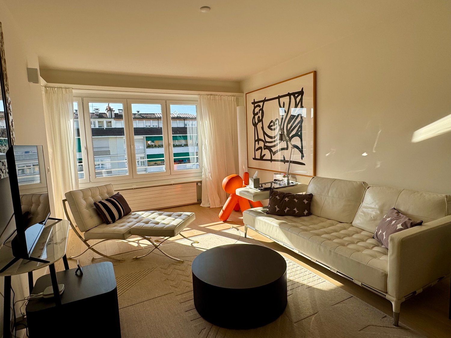 Spacious living room with large windows, beige sofa, tufted armchair, black coffee table, and a large abstract artwork on the wall